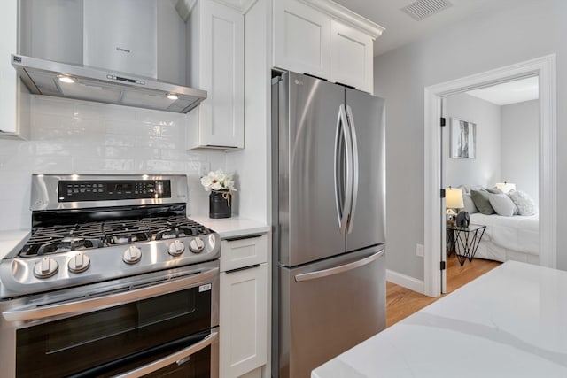 kitchen with tasteful backsplash, white cabinets, light hardwood / wood-style floors, wall chimney range hood, and appliances with stainless steel finishes