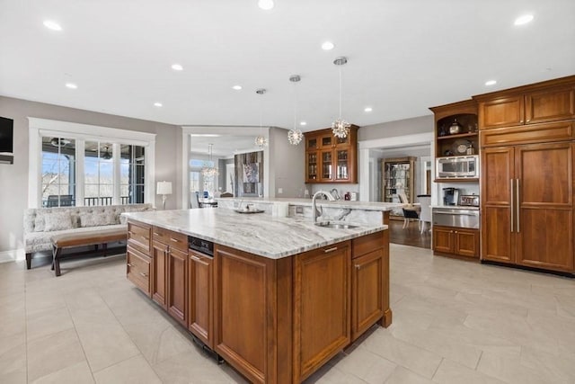 kitchen with light stone counters, sink, built in appliances, decorative light fixtures, and an island with sink