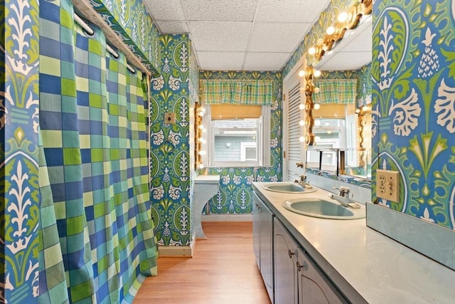 full bathroom with a sink, a drop ceiling, wallpapered walls, and wood finished floors