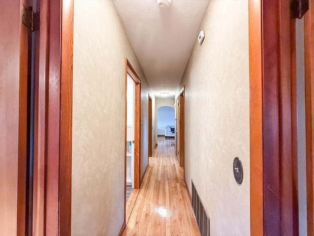 hall with light wood finished floors, visible vents, arched walkways, and a textured wall