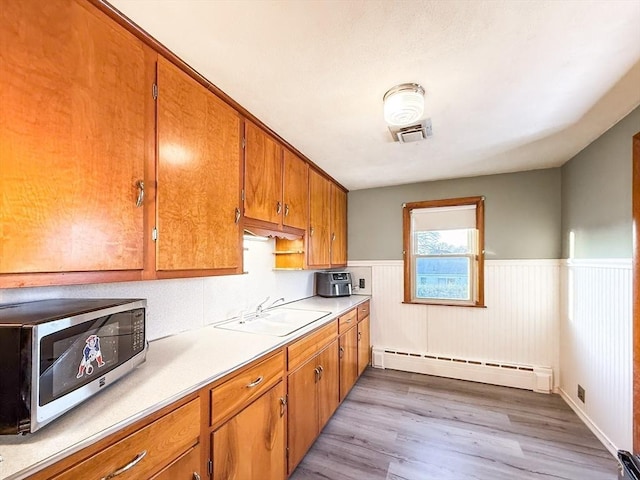 kitchen with brown cabinetry, light wood-style flooring, a sink, wainscoting, and baseboard heating