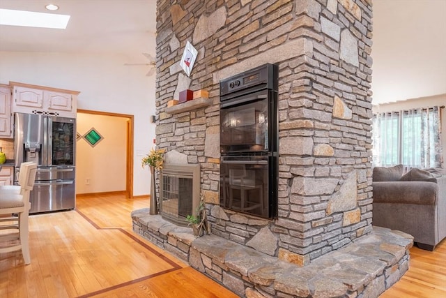 living room with a stone fireplace, a skylight, light wood-type flooring, baseboards, and high vaulted ceiling