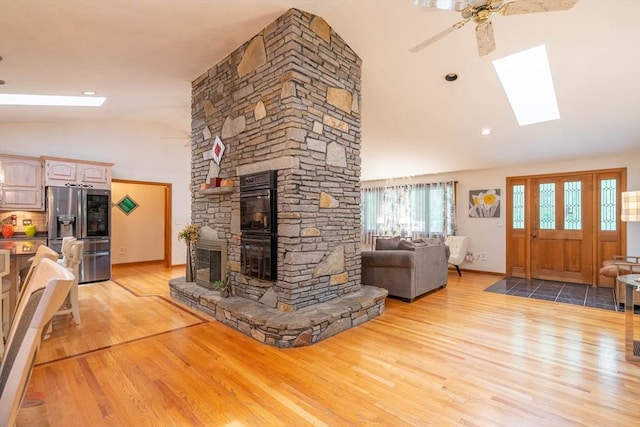 living area featuring a skylight, light wood-style flooring, ceiling fan, a stone fireplace, and high vaulted ceiling