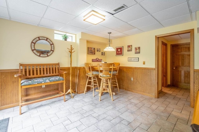 dining space with a drop ceiling, a wainscoted wall, wooden walls, a dry bar, and stone finish floors