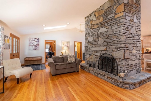 living room featuring vaulted ceiling, wood finished floors, a stone fireplace, a ceiling fan, and a skylight
