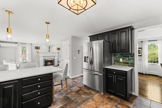 kitchen with stainless steel refrigerator with ice dispenser, dark cabinets, light countertops, a warm lit fireplace, and pendant lighting