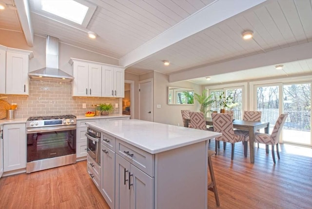 kitchen featuring light wood-style flooring, wall chimney exhaust hood, stainless steel appliances, light countertops, and backsplash