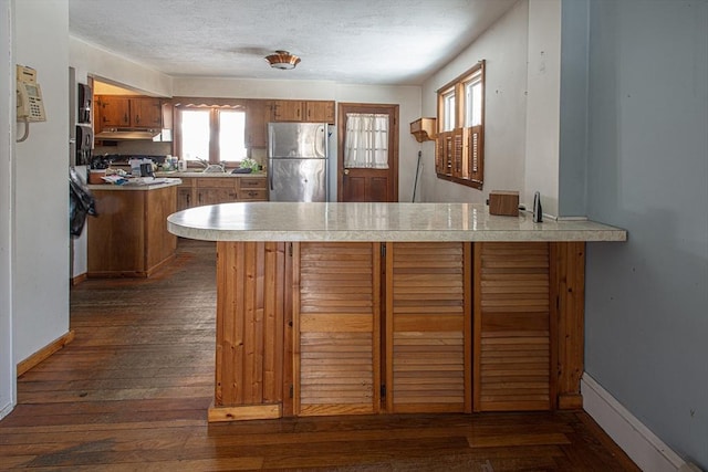 kitchen featuring light countertops, brown cabinetry, freestanding refrigerator, dark wood-type flooring, and a peninsula