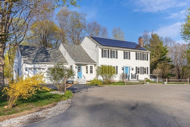 colonial home featuring a garage, a chimney, roof mounted solar panels, and aphalt driveway