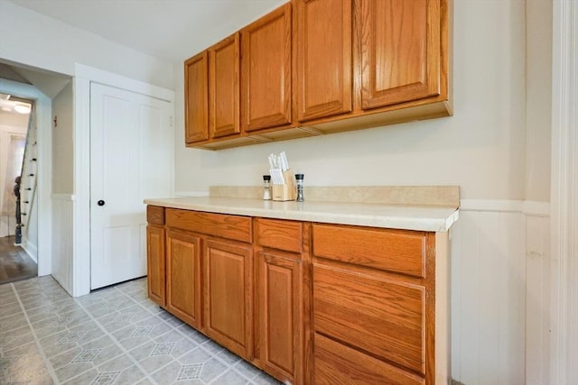 kitchen featuring wainscoting, light countertops, brown cabinetry, and a decorative wall