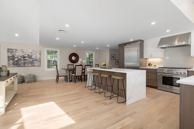 kitchen featuring high quality appliances, wall chimney range hood, a breakfast bar, a center island with sink, and light wood-style floors