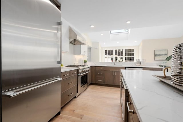 kitchen featuring premium appliances, wall chimney exhaust hood, light wood-style flooring, light stone counters, and dark brown cabinets