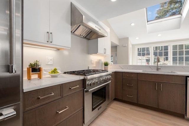 kitchen with premium appliances, wall chimney range hood, a sink, a skylight, and light wood-style flooring