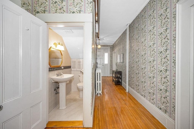 half bath with wallpapered walls, a sink, wood-type flooring, tile walls, and toilet