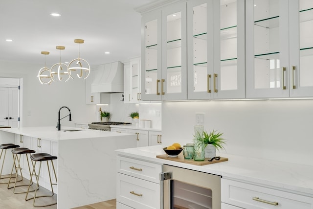 kitchen with a center island with sink, custom range hood, hanging light fixtures, sink, and white cabinets