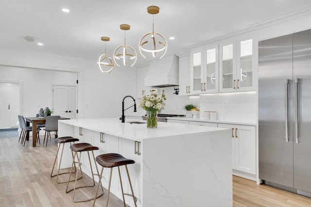 kitchen featuring built in refrigerator, premium range hood, an island with sink, and hanging light fixtures