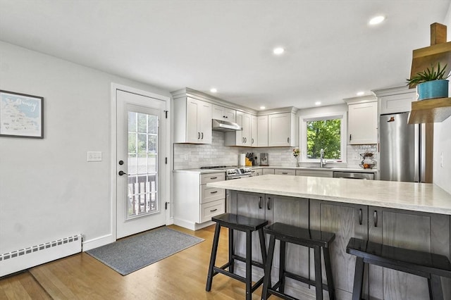kitchen featuring a baseboard heating unit, a kitchen bar, backsplash, appliances with stainless steel finishes, and under cabinet range hood