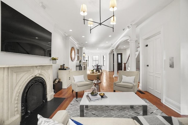 living area with a fireplace with flush hearth, wood finished floors, an inviting chandelier, crown molding, and ornate columns