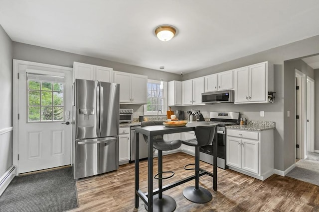 kitchen with stainless steel appliances, wood finished floors, a baseboard heating unit, white cabinets, and light stone countertops