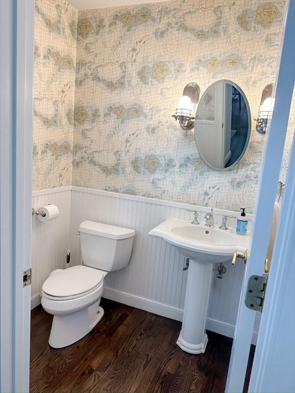 half bathroom with wood finished floors, toilet, wallpapered walls, and wainscoting