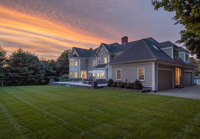 back of house at dusk with a garage, driveway, a shingled roof, a chimney, and a yard