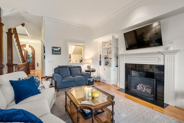 living room featuring ornamental molding, a tiled fireplace, built in shelves, wood finished floors, and stairway