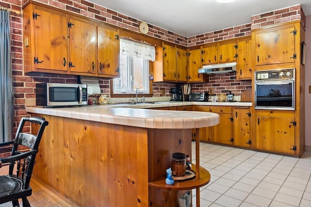 kitchen featuring sink, oven, tile countertops, kitchen peninsula, and brick wall