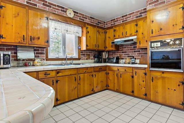 kitchen with sink, tile countertops, black oven, light tile patterned floors, and brick wall