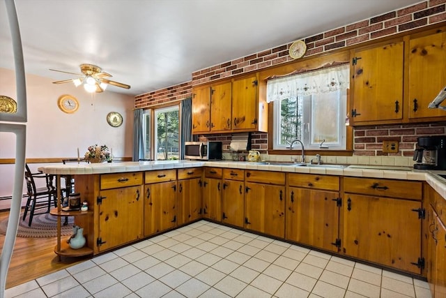 kitchen featuring sink, tile countertops, ceiling fan, light tile patterned floors, and brick wall