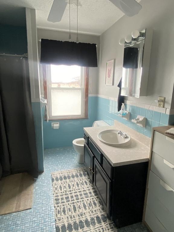 bathroom with a ceiling fan, a wainscoted wall, a textured ceiling, vanity, and tile walls