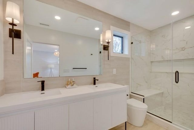 full bath featuring tile walls, a marble finish shower, double vanity, toilet, and a sink