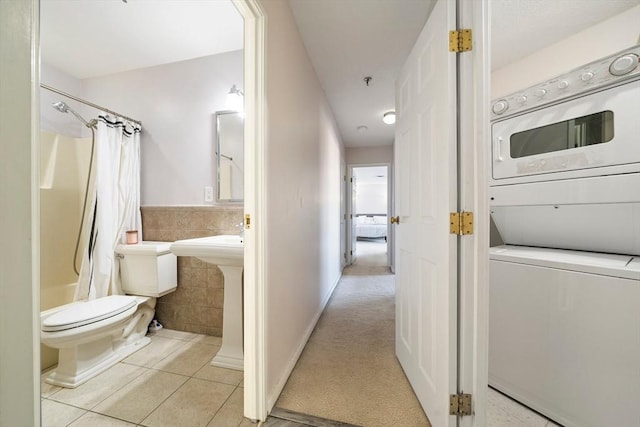 bathroom featuring tile walls, toilet, shower / bath combo with shower curtain, stacked washer / dryer, and tile patterned flooring