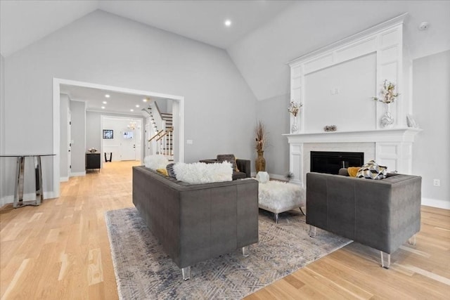 living area featuring stairway, baseboards, high vaulted ceiling, light wood-style floors, and a glass covered fireplace