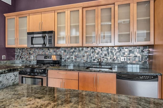 kitchen with dark stone countertops, a sink, glass insert cabinets, appliances with stainless steel finishes, and tasteful backsplash