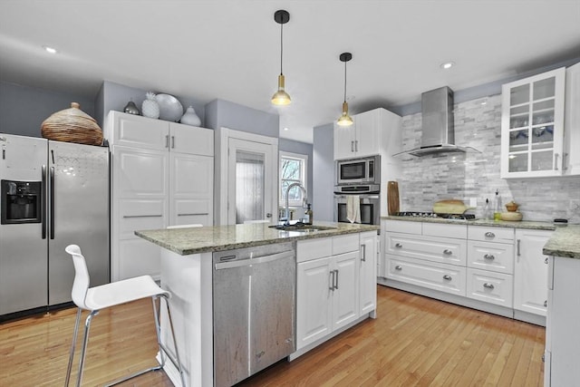 kitchen with wall chimney range hood, appliances with stainless steel finishes, light wood-style floors, white cabinetry, and a sink