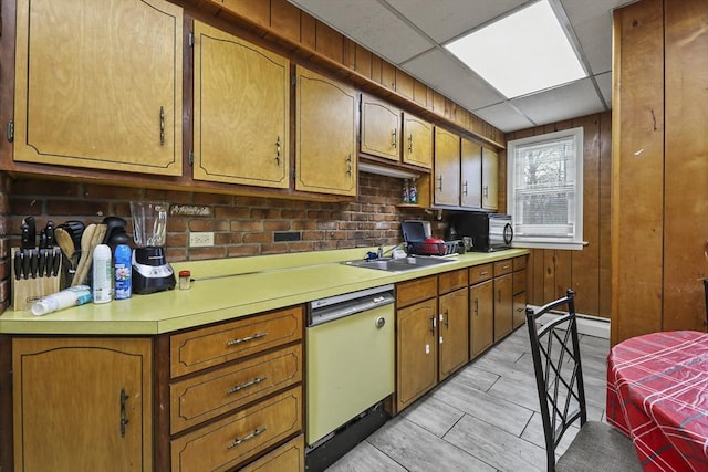 kitchen with a sink, wood walls, light countertops, dishwashing machine, and a paneled ceiling