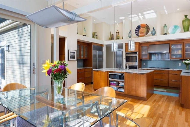 kitchen with appliances with stainless steel finishes, decorative light fixtures, backsplash, a kitchen island with sink, and wall chimney range hood