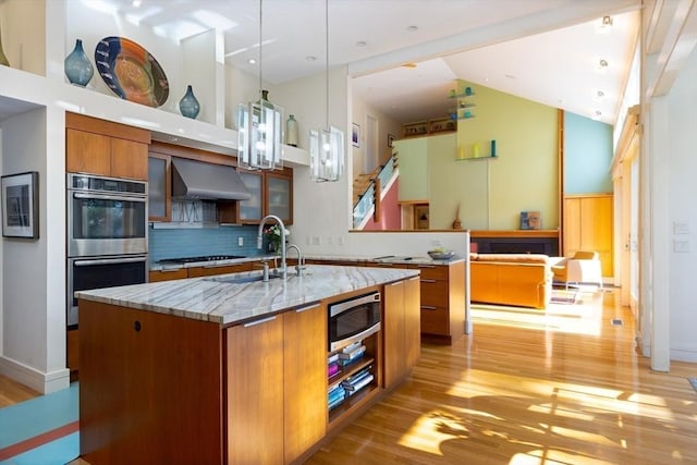 kitchen with decorative backsplash, hanging light fixtures, wall chimney range hood, stainless steel appliances, and a center island with sink