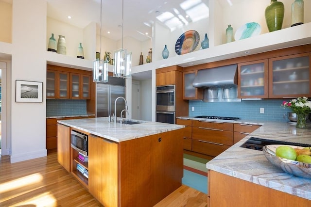 kitchen with range hood, an island with sink, sink, decorative backsplash, and built in appliances