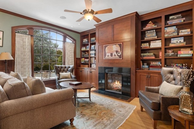 sitting room with built in shelves, a fireplace with flush hearth, ornamental molding, light wood-style floors, and a ceiling fan