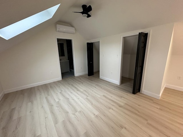 unfurnished bedroom featuring vaulted ceiling with skylight, a wall mounted air conditioner, ensuite bath, ceiling fan, and light wood-type flooring