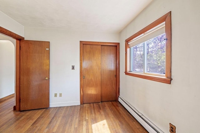 unfurnished bedroom with a baseboard heating unit, wood-type flooring, a closet, baseboards, and arched walkways