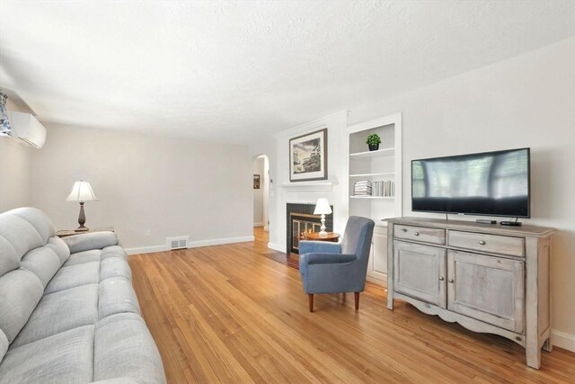 living area featuring light wood finished floors, an AC wall unit, built in features, a fireplace with flush hearth, and a textured ceiling