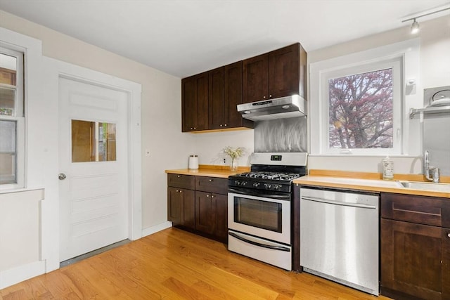 kitchen featuring dark brown cabinets, a sink, under cabinet range hood, light wood-style floors, and stainless steel appliances