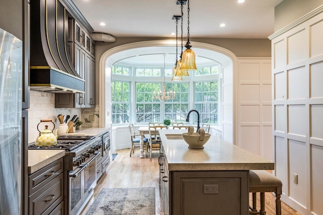 kitchen featuring custom exhaust hood, double oven range, backsplash, light wood-type flooring, and a kitchen island with sink