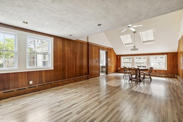 unfurnished dining area featuring ceiling fan, lofted ceiling with skylight, light hardwood / wood-style floors, wooden walls, and baseboard heating