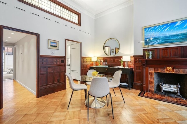 dining room with a towering ceiling, a tile fireplace, crown molding, and a wainscoted wall