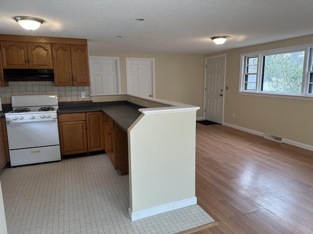 kitchen featuring brown cabinetry, dark countertops, under cabinet range hood, and gas range gas stove