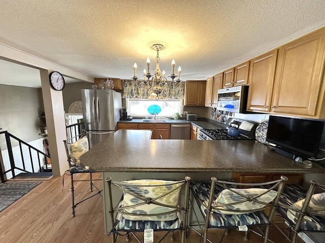 kitchen featuring wood finished floors, an inviting chandelier, a peninsula, stainless steel appliances, and dark countertops