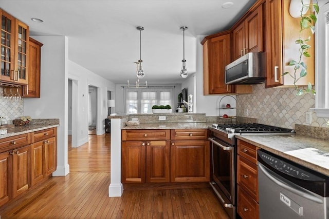 kitchen featuring decorative light fixtures, appliances with stainless steel finishes, light stone countertops, brown cabinetry, and glass insert cabinets